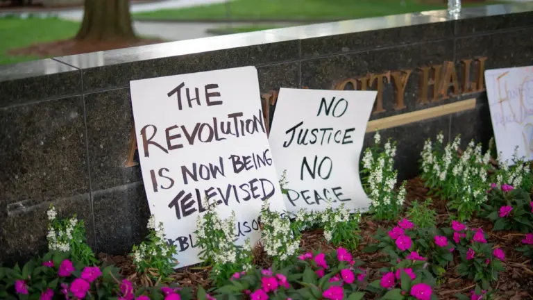 Pancartes de protestation posées devant un mur en pierre avec des fleurs roses.